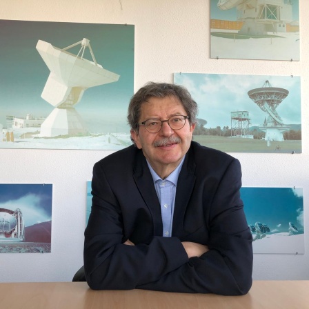 Karl Menten trägt eine Brille und sitzt an einem Schreibtisch. Hinter ihm sind Fotos von Satellitenanlagen zu sehen.