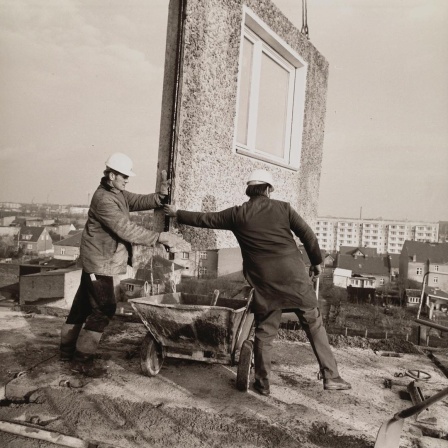 Eine Schwarz-Weiß-Aufnahme aus den 1970er Jahren zeigt Bauarbeiter beim Einfügen eines Teilstücks für ein Wohnhaus, das in Plattenbauweise errichtet wird.