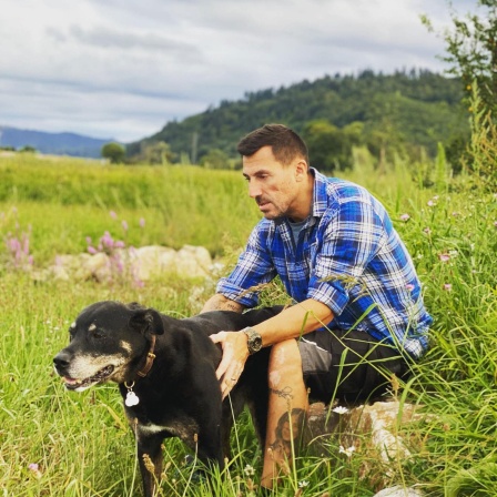 Christian Schindler sitzt auf einem großen Stein auf einer Wiese. Vor sich ein schwarzer Hund, den er streichelt.