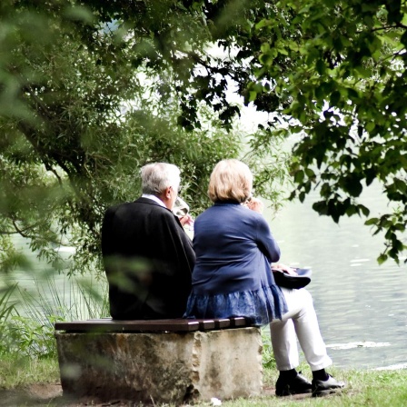 Ein älteres Ehepaar sitzt auf einer Bank vor einem See und trinkt Wein.