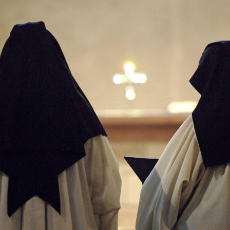 Zwei Zisterzienserinnen stehen ein einem Kloster in Deutschland bei der Messe mit dem Rücken zur Kamera. Sie tragen ein weißes Gewand und einen schwarzen Nonnenschleier.