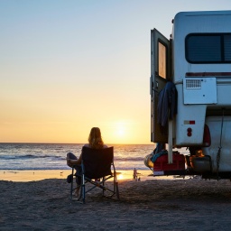 Eine Frau sitzt bei Sonnenuntergang neben einem Wohnmobil am Strand