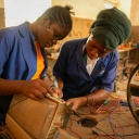 Zwei Schwarze Frauen arbeiten an einem Werkstück aus Holz. Um sie herum liegen Kabel.