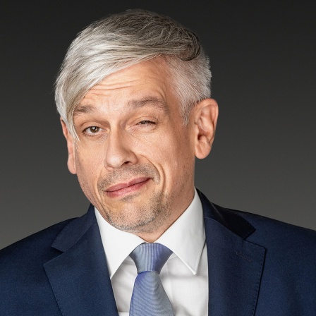 Der Kabarettist Mathias Tretter mit Anzug vor dunklem Hintergrund zwinkert in ide Kamera