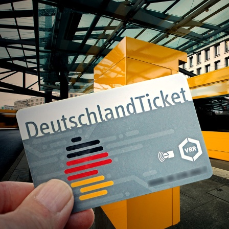 Eine Hand hält ein Deutschlandticket, im Hintergrund fährt eine Straßenbahn an einer Haltestelle