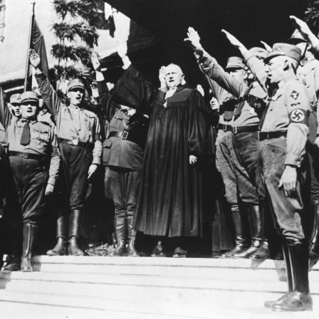Schwarzweißfoto zeigt einen Mann in Talar, den Hitlergruß zeigend vor einem Spalier von SA-Uniformierten mit erhobenem rechten Armen.