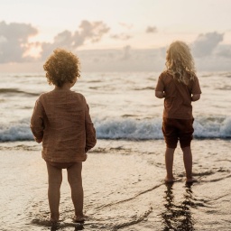 Zwei Kinder stehen knöchelhoch im Wasser an einem Strand bei Sonnenuntergang.
