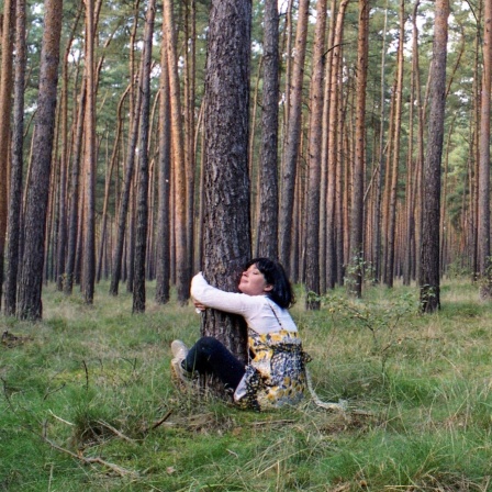 Eine Frau umarmt im Wald sitzend einen Baum.