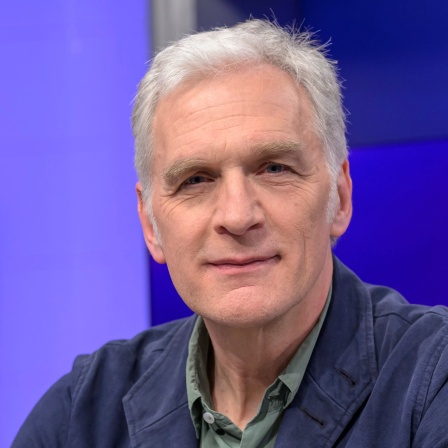 Der Schauspieler Walter Sittler sitzt vor einer blauen Wand und schaut in die Kamera.