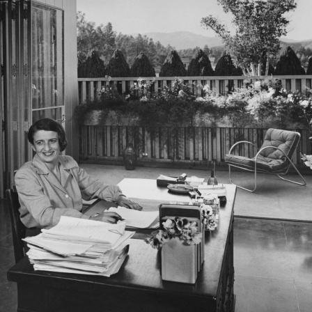 Die Schriftstellerin Ayn Rand sitzt an ihrem Schreibtisch in ihrem von Richard J. Neutra entworfenen Haus im San-Fernando-Tal; sie lächelt und schaut mit den Augen von der Kamera weg; sie trägt eine grau geknöpfte Bluse und hat beide Hände auf dem Sch