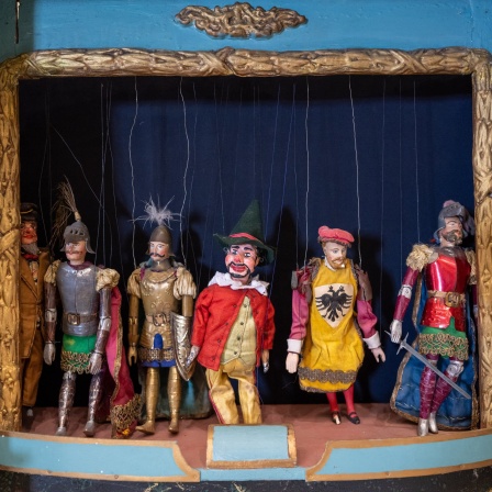 Ein historisches Puppentheater von circa 1870 steht im Münchner Marionettentheater neben der Bühne: Marionetten erscheinen auf der Bühne u. a. deshalb so groß, weil die Requisiten - also die Gegenstände, mit denen sie zu tun haben, wie Stühle, Tische oder Türen - im Verhältnis etwas kleiner gestaltet sind.