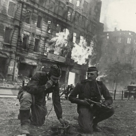 Soldaten der Roten Armee knien vor einem brennenden Haus in Berlin. Einer hält einen Hörer ans Ohr, der andere hat ein Maschinengewehr in der Hand.  