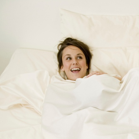 Ein Paar im Bett, die Frau lacht während der Mann unter der Decke verschwindet.