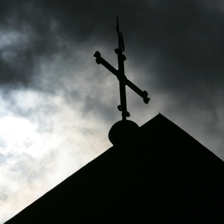 Im Gegenlicht und vor wolkenverhangenem Himmel ist eine Kirchturmspitze mit Kreuz zu sehen
