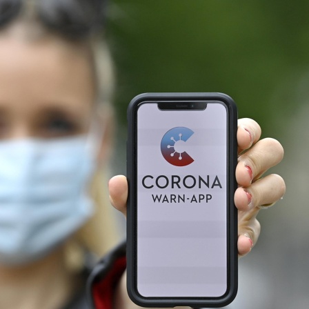 Ein Handy mit der geöffneten Corona Warn-App wird von einer Frau in die Luft gehalten.