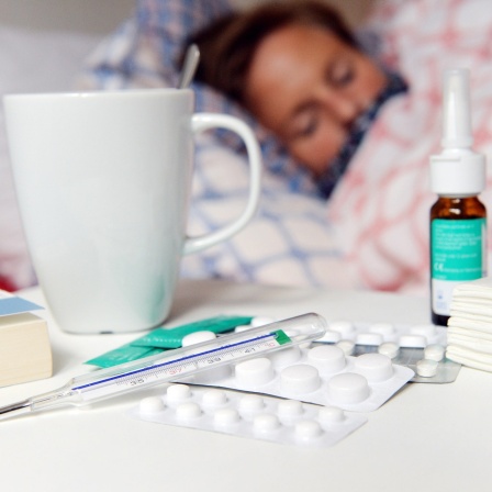 Medikamente und ein Fieberthermometer liegen auf einem Nachttisch vor einer Frau, die im Bett liegt.