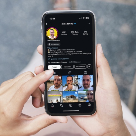 Eine Frau hält ein Smartphone in den Händen auf dem das Instagram-Profil eines Immobilien-Influcencers zu sehen ist.