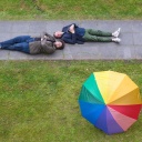 Devid Striesow und Axel Rahnisch liegen rücklings auf einem Fußgängerweg durch eine grüne Wiese, auf der widerum ein regenbogenfarbener Schirm liegt.