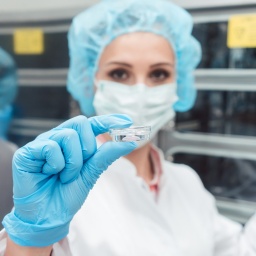 Eine Wissenschaftlerin im Laborkittel mit Mundschutz, Gummihandschuhen und Haarnetz hält ein Glasgefäß hoch.