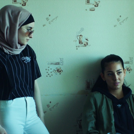 Eine Filmszene aus der Netflix-Serie Kalifat zeigt zwei Frauen. Eine trägt ein Kopftuch und steht, die zeit hockt an einer grünen Wand.