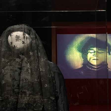 Schneiderpuppe, vor einer Projektion eines Mannes, welche einen schwarzen Schleier über das dem Gesicht trägt