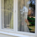 Eine ältere Frau steht hinter der Gardine am Fenster. 