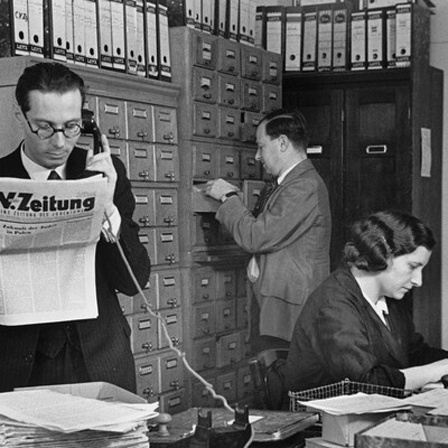 Zwei Männer und eine Frau in der Redaktion der C.V.- Zeitung. Die Frau am Schreibtisch sitzend, hinten ein Mann an einem Regal, vorne Hans (John F.) Oppenheimer mit Telefonhörer und C.V.- Zeitung in den Händen.