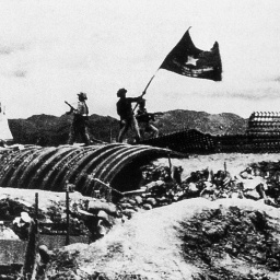 Eine historische Schwarzweiß-Aufnahme zeigt das Schlachtfeld von Dien Bien Phu in Vietnam. Ein Soldat hält die vietnamesische Flagge hoch.