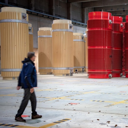 Eine Person läuft durch eine Halle, in der reihenweise tonnenförmige Behälter in beige und rot stehen, die Atommüll beinhalten.