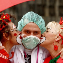 Mann mit Mütze und Mundschutz wird von zwei Frauen in Clownkostüm geküsst