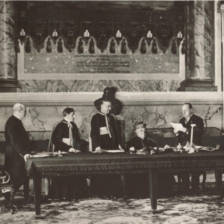 Ein Schwarz-Weiß-Foto zeigt die Vertragsunterzeichnung im Lateranspalast am 11. Februar 1929 mit Kardinalstaatssekretär Pietro Gasparri und dem faschistischen Ministerpräsidenten Italiens Benito Mussolini am Mikrofon stehend.
