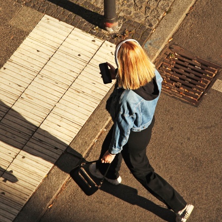 Eine junge Frau trägt Kopfhörer und blickt auf ihr Smartphone, während sie über eine Straße geht