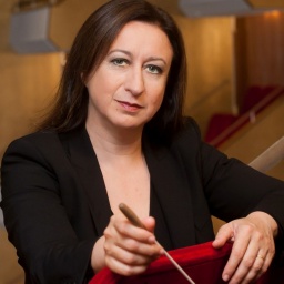 Eine Frau sitzt verkehrt herum auf einem roten Sessel und hält einen Dirigentenstab in der Hand, während ihr Blick ernst ist. 