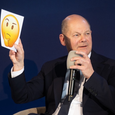 Bundeskanzler Scholz hält in einer Podiumsdiskussion einen Zettel mit einem nachdenklich guckenden Emoji nach oben.