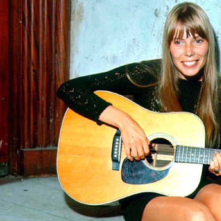 Joni Mitchell sitzt mit ihrer Gitarre vor einer Holztür und lächelt