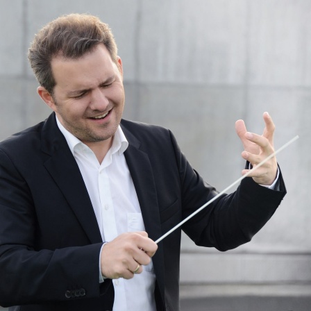 Mann mit schwarzem Sakko und weißem Hemd und Taktstock, Dirigentengeste mit der linken Hand, konzentriert-fröhlichen nach unten blickend