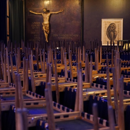 Die Stühle in einer Kirche sind allesamt hochgestellt, im Hintergrund ist eine Jesus-Figur an der Wand zu sehen.