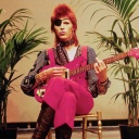 David Bowie sitzt auf einem Stuhl und hält eine E-Gitarre in der Hand, daneben ein leerer Vogelkäfig.