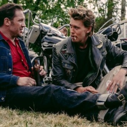 Im Still aus "The Bikeriders" sitzen Tom Hardy und Austin Butler nebeneinander im Rasen an ihre Motorräder gelehnt und reden.