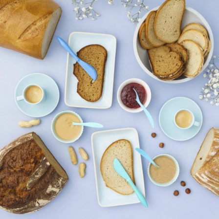 Frühstück auf einem hellen Tisch mit verschiedenen Brotsorten, Aufstrich und Erdnussbutter.