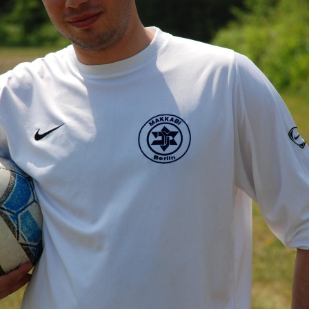 Ein Spieler des Vereins TuS Makkabi Berlin trägt ein weißes Trikot des Vereins, unter dem rechten Arm hat er einen Fußball geklemmt. 