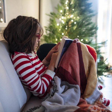 Ein Kind sitzt im Schlafanzug auf dem Sofa vor dem Christbaum und liest in einem Buch.