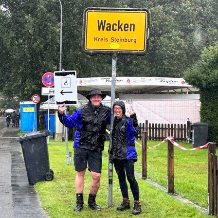 Zwei Wacken-Besucher stehen unter dem Ortschild mit der Aufschrift "Wacken" und schauen in Richtung Kamera