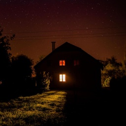 Nachts: Blick auf ein einsames Haus auf einer Wiese, am Rand Bäume.