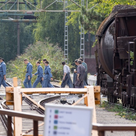 Statisten gehen 2022 im Rahmen der Dreharbeiten zum Film "Die Tribute von Panem: Das Lied von Vogel und Schlange" im öffentlichen Teil des Landschaftsparks am Erkennungszeichen des Parks vorbei, einer alten Lok