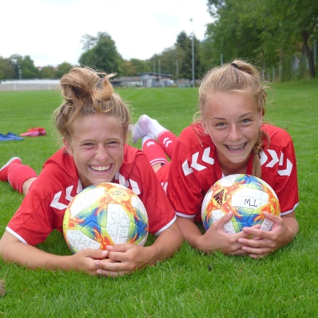 zwei junge Fußballspielerinnen liegen im Gras und lachen in die Kamera