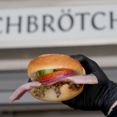 Ein Mitarbeiter hält ein Fischbrötchen mit Matjes vor dem Schriftzug "Fischbrötchen" an einem Imbiss an den Landungsbrücken im Hafen hoch