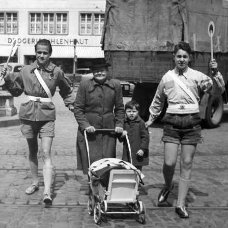 Zwei Schülerlotsen haben eine Frau mit Kinderwagen und einem kleinen Jungen in die Mitte genommen und führen sie mit erhobenen Kellen über eine Straße (Aufahme von 1953)