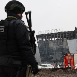 Ein Polizeibeamter patrouilliert am 23. März 2024 am Ort des Schusswaffenangriffs in der Konzerthalle „Crocus City Hall“ in Krasnogorsk bei Moskau. Im Vordergruns ist der Polizist in voller Montur zu sehen, im Hintergrund die abgebrannte Stadthalle.