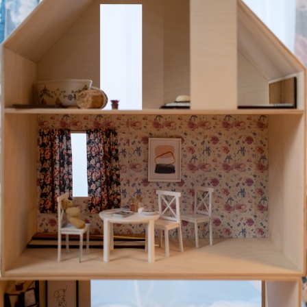 Blick ins Zimmer eines Holzpuppenhauses mit einem runden, weißen Esszimmertisch und drei Stühlen, vor einer Wand mit Blumentapete. Auf einem Stuhl sitzt ein Teddybär.